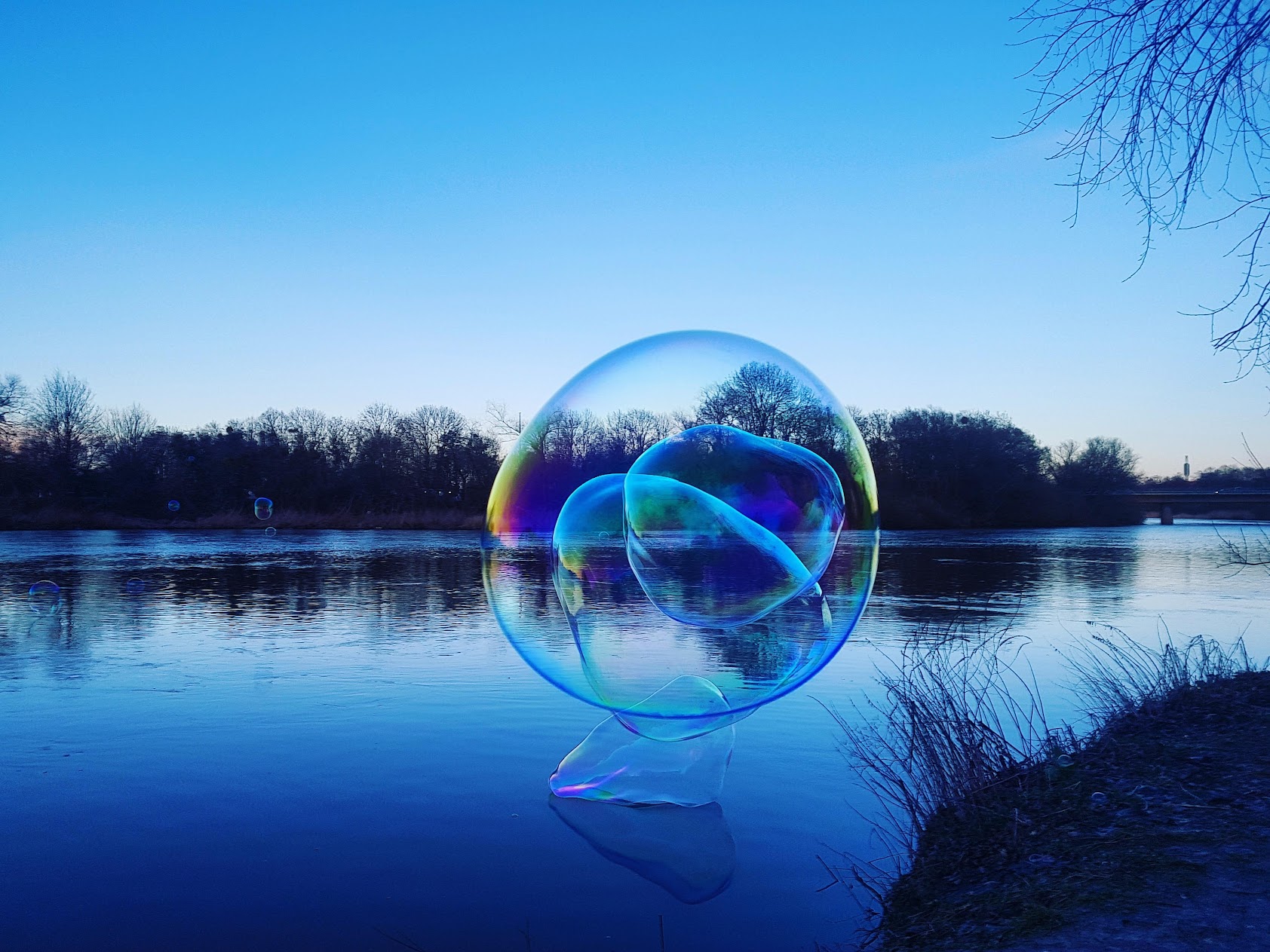 Eine schimmernde Riesenseifenblase schwebt zur blauen Stunde &uuml;ber einem Fluss