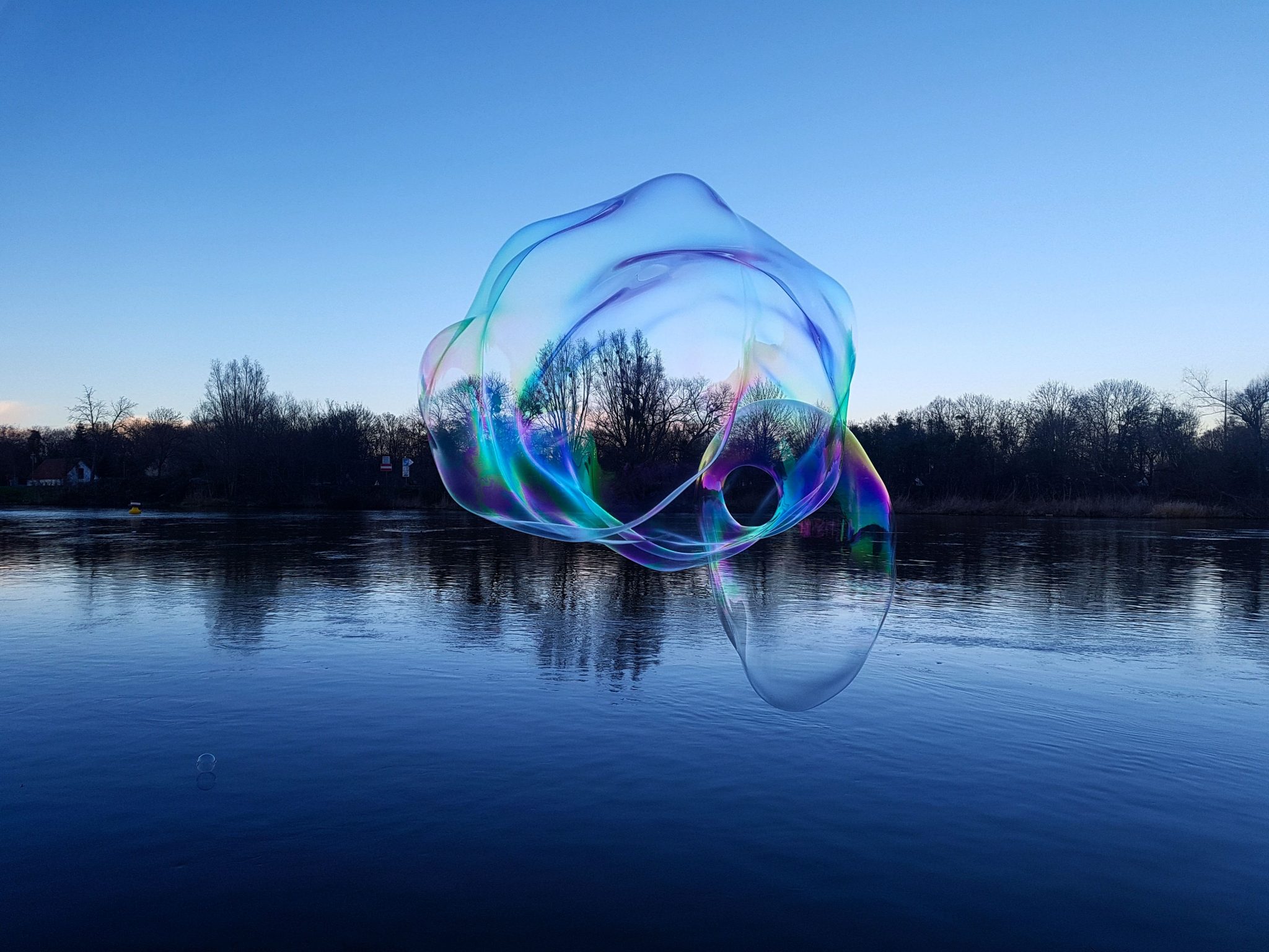 Eine riesige schillernde Seifenblase schwebt zur blauen Stunde &uuml;ber einem spiegelglatten See