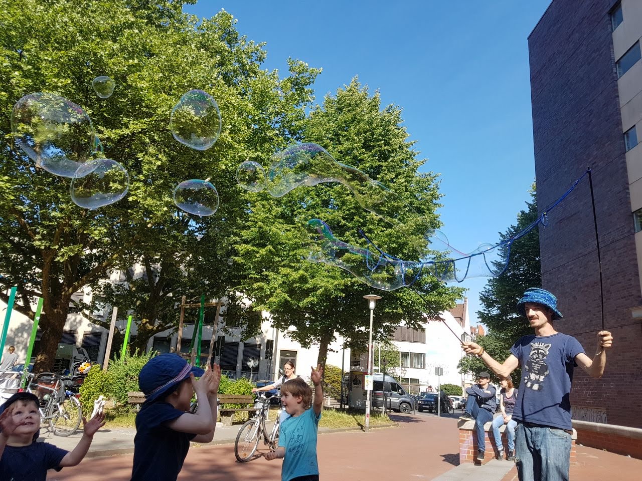 Roger macht auf einem Stadtplatz Seifenblasen, denen Kinder mit erhobenen H&auml;nden nachjagen